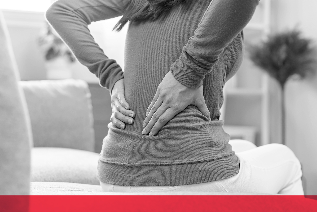 Black-and-white photo of a woman sitting and holding her lower back with both hands, indicating back pain