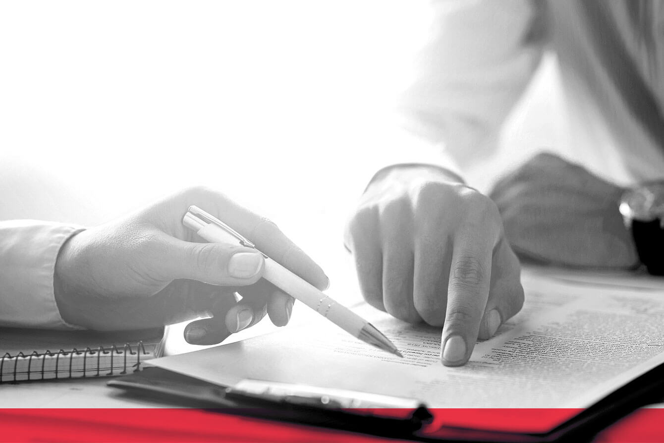 A close-up of two people reviewing a document. One person is holding a pen, while the other is pointing at a specific section of the text. A notepad and a clipboard are visible on the desk. The image is in black and white with a red accent at the bottom.